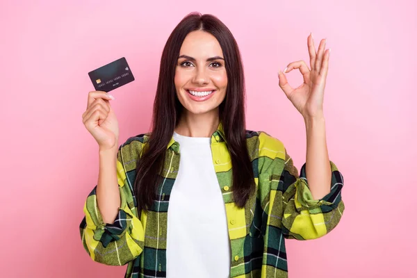 Portrait of satisfied lovely person long hairstyle yellow jacket hold credit card show okey approve isolated on pink color background.