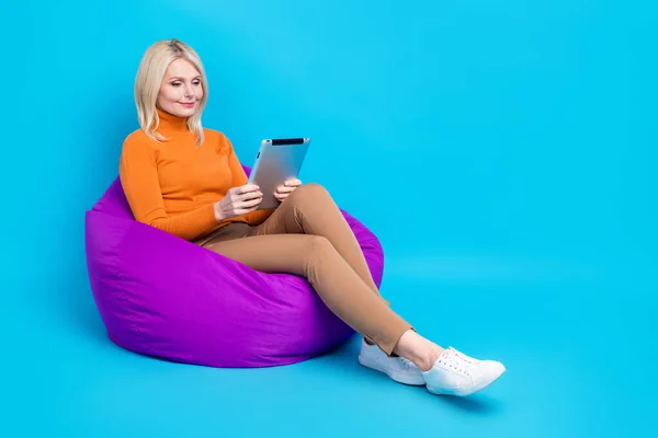 満足した平和的な女性の完全な長さの写真は オレンジ色のスウェットシャツを身に着けている豆の袋の上に座って読むEbook隔離された青い色の背景 — ストック写真