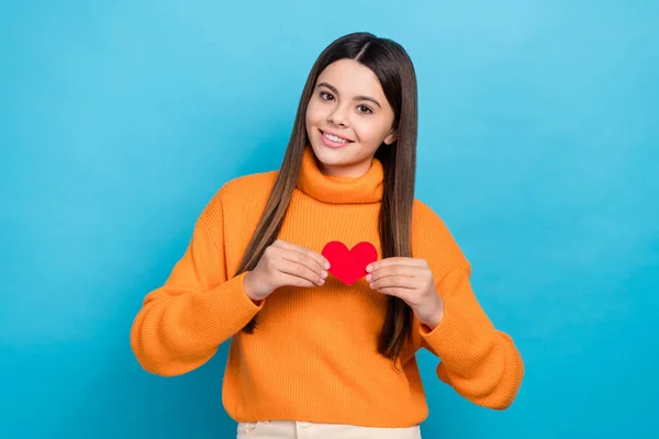 Photo of satisfied kind cute girl with straight hairdo wear orange sweater holding small paper heart isolated on blue color background.