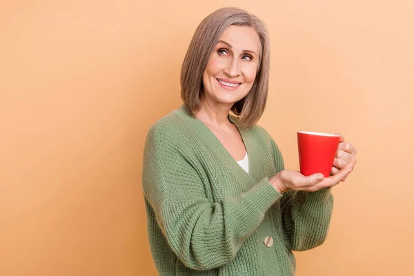 Photo of dreamy grandmother wear trendy khaki shirt hold cup tea enjoy chinese brand look empty space isolated on beige color background.