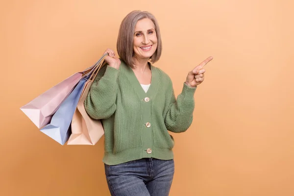 友好的祖母穿着针织套头衫 拿着购物袋 向顾客提供用彩绘背景隔开的空旷空间 — 图库照片