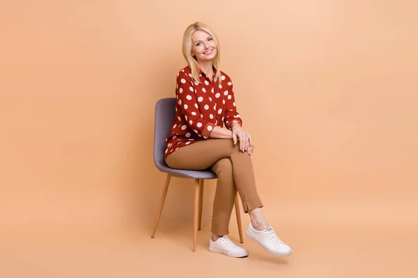 楽観的な先輩女性の完全な長さの写真ドレス赤いシャツパンツ座っています上の椅子にインタビューで隔離されたベージュ色の背景 — ストック写真