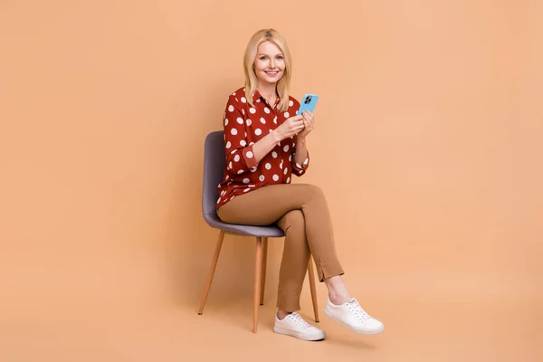 正の女性座る椅子の完全な長さの肖像画使用スマートフォンチャット絶縁でベージュ色の背景 — ストック写真