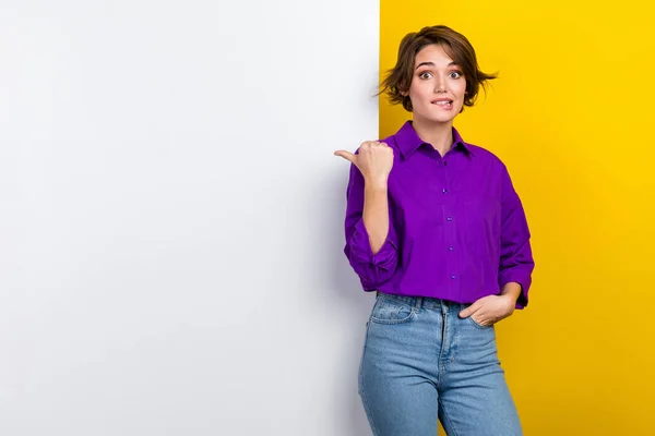 年轻的营销小姐穿着紫色衬衫咬嘴唇的照片令人怀疑的指向黄色背景的手指纸广告牌 — 图库照片