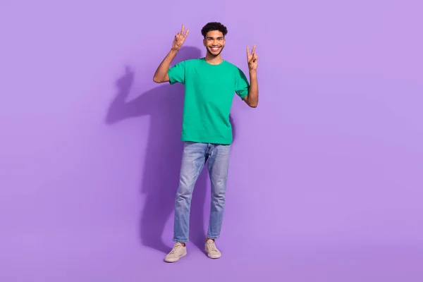 若い男の挨拶のシンボルの完全な長さの写真Vサインこんにちはは 紫の背景に隔離された緑のTシャツデニムカートジーンズを身に着けている — ストック写真