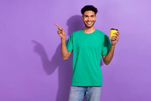 照片上积极乐观的小伙子身穿绿色T恤 享受可可 指向空旷的空间 孤立的紫色背景 — 图库照片
