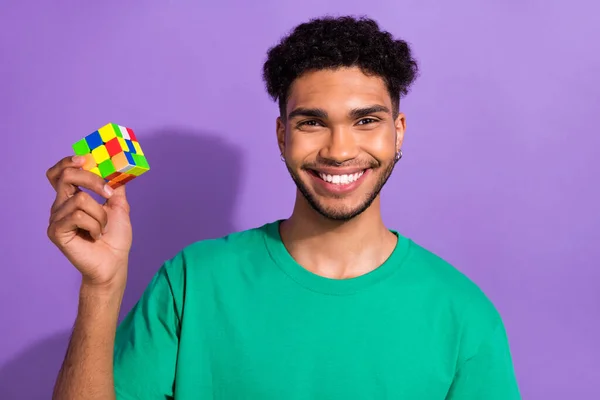 Photo of cool good mood man wear green t-shirt smiling rising rubik cube isolated purple color background.