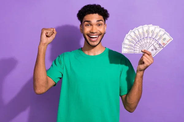 Photo of funky lucky man wear green t-shirt rising fist showing money fan isolated purple color background.