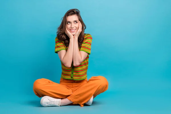 Full size photo of minded woman dressed orange pants colorful cardigan look empty space arms cheekbones isolated on blue color background.