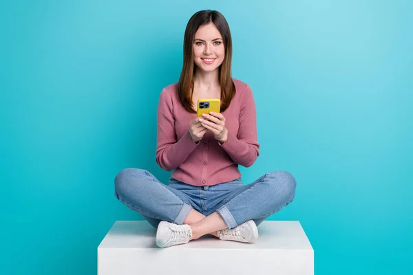 フルサイズの写真の良い気分かわいい女の子服ピンクカーディガンジーンズホールドスマートフォン座って上のキューブに隔離された青色の背景 — ストック写真