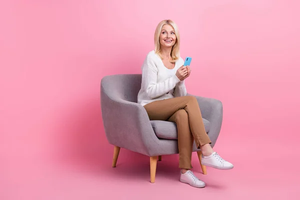 フルサイズの肖像画の素敵な正の人座って快適椅子使用スマートフォンは空のスペースに見えるピンク色の背景に隔離 — ストック写真