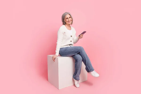 フルボディ写真の豪華な成熟したおばあちゃん座ってキューブ電話を保持ドレススタイリッシュな白い服ピンク色の背景に隔離 — ストック写真