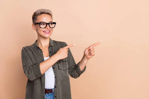 Photo of young cheerful lady show blonde haircut khaki shirt direct fingers look empty space store isolated on beige color background.