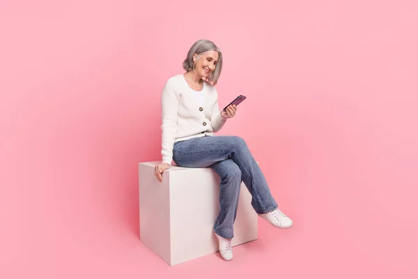 魅力的なシニア女性のフルサイズの写真座ってキューブホールドデバイス読み取りニュースピンク色の背景に隔離された流行の白い服を着用 — ストック写真