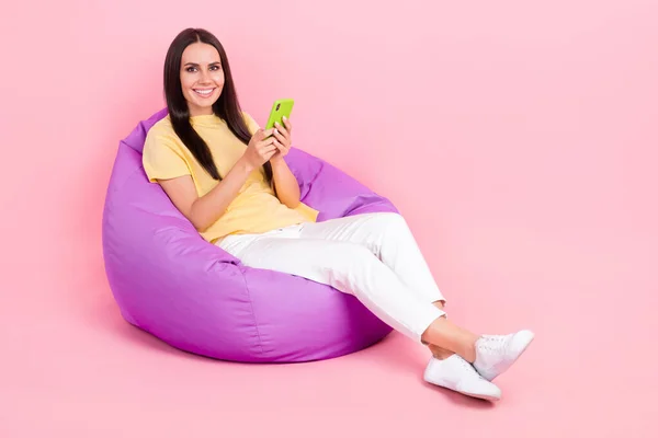 フル長さの写真正の女性の服黄色Tシャツ座って豆袋タイピングSmsデバイス空のスペース孤立ピンク色背景 — ストック写真