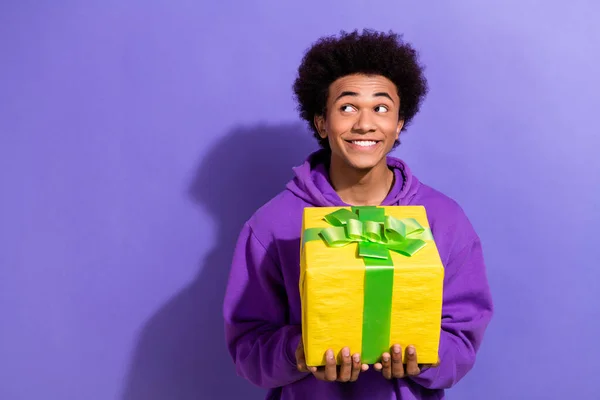 Portrait of positive minded man toothy smile arms hold giftbox look interested empty space isolated on purple color background.
