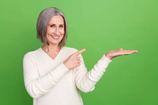 有魅力的退休女性推销商的照片直接指向她张开的手掌 展示新奇的广告广告和产品孤立的绿色背景 — 图库照片