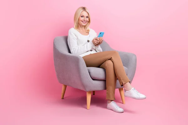 良い気分人の完全な長さの写真ピンク色の背景に隔離されたスマートフォンを入力アームチェアに座って白いセーターを身に着けている — ストック写真