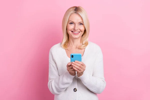 明るい正の満足シニア女性の写真ニットカーディガンホールドスマートフォンのタイピングポストピンク色の背景に隔離された — ストック写真