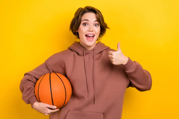 满意的漂亮女子身穿褐色帽衫的照片 就像拿着黄色背景的篮球 — 图库照片