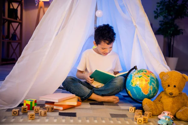 stock image photo of positive clever geek nerd boy sit in homemade tent read interesting book in evening playroom indoors.