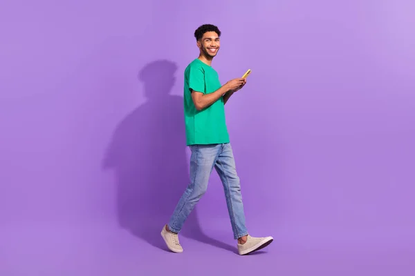若い男の完全な長さの幹部は 紫の色の背景に隔離されたスマートフォンのオンラインストアショッピングブラウザで緑のTシャツの歩行を着用 — ストック写真
