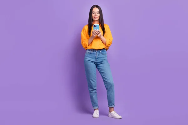 若い自信のあるビジネス女性の完全な長さの写真は オレンジ色のシャツデニムジーンズを身に着けている読み取り電子メールホールド電話絶縁紫の色の背景 — ストック写真