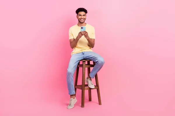 年轻的心满意足的男人坐在椅子上的全副身体照片 用智能手机浏览被粉色背景隔离的Safari互联网页面 — 图库照片