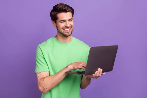 穿着绿色T恤衫的聪明而积极的男人的画像看着手提电脑 手里拿着用紫色背景隔开的电子邮件打字 — 图库照片