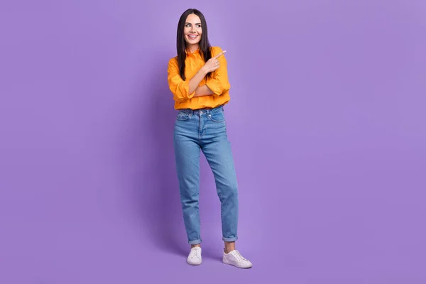 笑顔の女の子のジーンズのフルボディ写真は オレンジ色のシャツを着用直接指は紫色の背景に隔離された空のスペースの商業プロジェクトに見えます — ストック写真