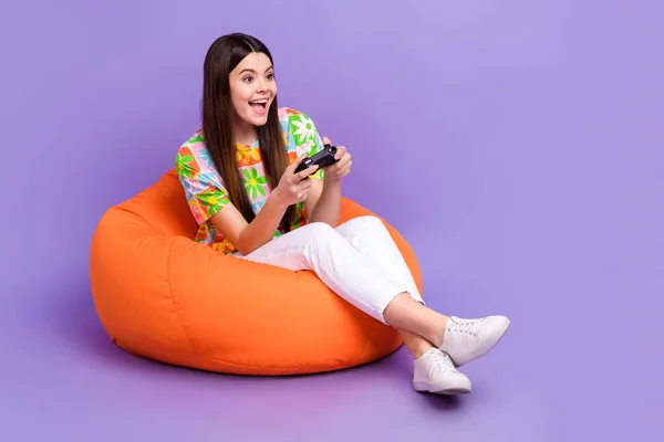 Full size photo of satisfied person wear stylish t-shirt sit on pouf hold joystick look empty space isolated on purple color background.