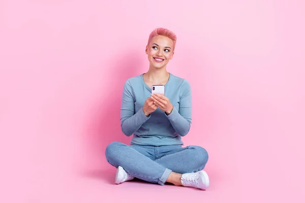 フレンドリーな女性のフルサイズの写真は スマートフォンで床に座ってスタイリッシュなジーンズを着用します ピンク色の背景に隔離された空のスペース — ストック写真