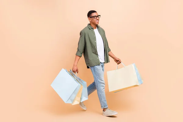 Full length profile photo of positive man hold mall bags walking empty space isolated on beige color background.