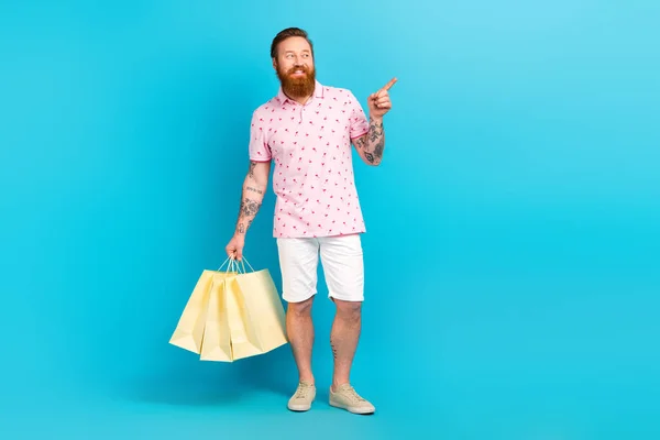 Full body portrait of eccentric positive man hold mall bags look direct finger empty space isolated on blue color background.