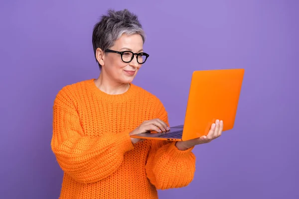 老退休妇女橙色套头衫手拿着新的上网本 集中工作的边远自由职业者 背景为紫罗兰色 — 图库照片