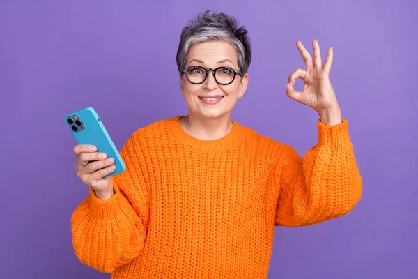 甜美而时髦的女人穿着橙色毛衣发短信现代装置的照片 照片上有一个迷人的手势 孤立的紫色背景 — 图库照片
