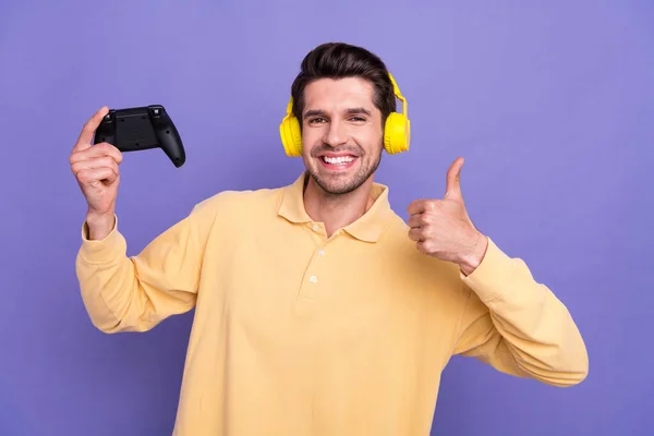 クールな自信を持った男の写真 黄色いシャツのイヤホンを着用 親指を上げる プレイステーションゲーム 孤立したヴァイオレット色の背景 — ストック写真