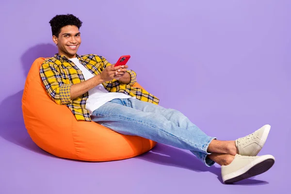 Full body portrait of positive funky person sit comfort bag use smart phone chatting isolated on purple color background.