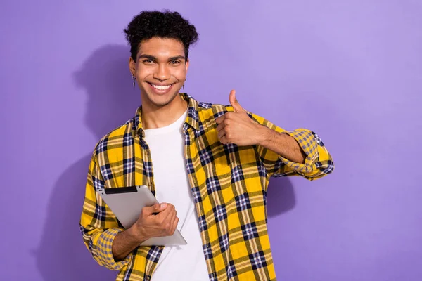 Photo of good mood cool guy dressed checkered yellow shirt reading modern gadget thumb up empty space isolated purple color background.