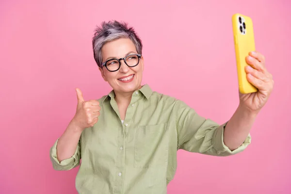 照片中快乐友善的资深女商人大拇指向上竖起 自拍干部相机穿着卡其布风格的衬衫 衬托着粉色的背景 — 图库照片