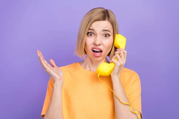 照片中 一个穿着黄色T恤 头型蓬松 在固话电话中与紫色背景隔离的小女孩惊讶地凝视着 — 图库照片