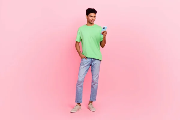 巧妙な陽気な男のフルサイズの写真 緑のTシャツのジーンズ スマートフォンを見て ピンク色の背景で隔離された電子メールを読む — ストック写真