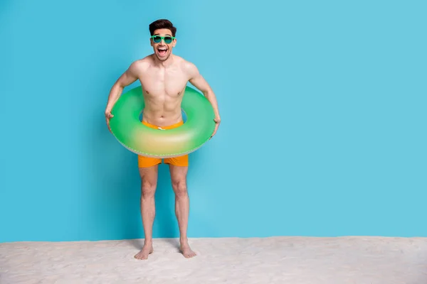 Full body photo of handsome young guy inflatable ring excited have swim dressed stylish yellow shorts isolated on blue color background.