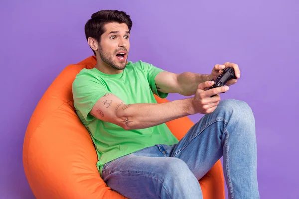 Photo of young funny staring man hold playstation gamepad console sitting pouf addicted losing isolated on violet color background.