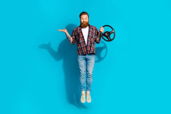 Full body photo of positive man dressed checkered shirt jeans palm presenting offer hold steering wheel isolated on blue color background.