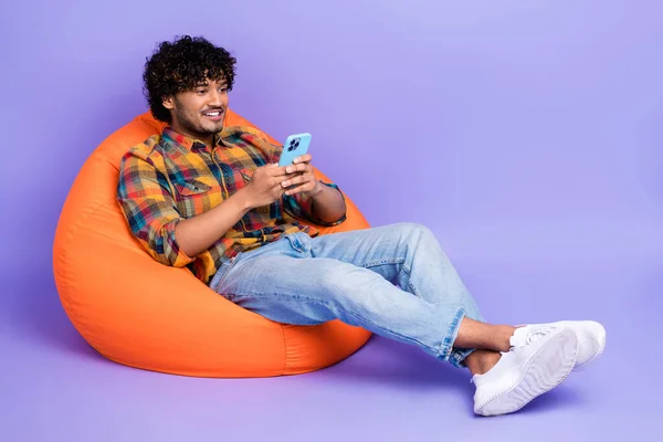 全长照片 英俊男子坐在橙色舒适豆袋上看智能手机 用紫色背景隔离留言 — 图库照片