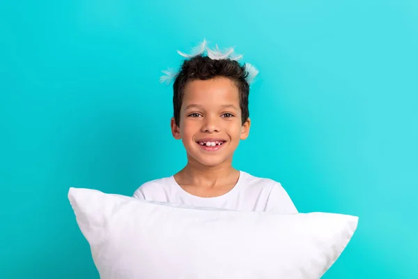 ターコイズ色の背景に隔離された微笑みの枕クッションフェザーヘッドをビームにしている陽気な小さな少年の肖像画 — ストック写真