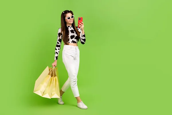 Full body profile portrait of beautiful teen girl hold boutique bags use smart phone walk empty space isolated on green color background.