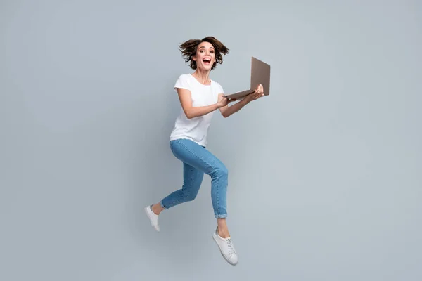 Full length photo of cute woman wear stylish t-shirt denim pants hold laptop hurry run in empty space isolated on gray color background.