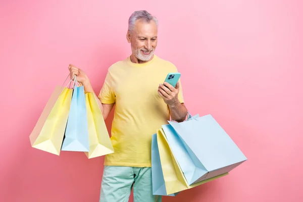 Portrait of satisfied person wear stylish t-shirt look at smartphone order taxi hold shopping bags isolated on pink color background.